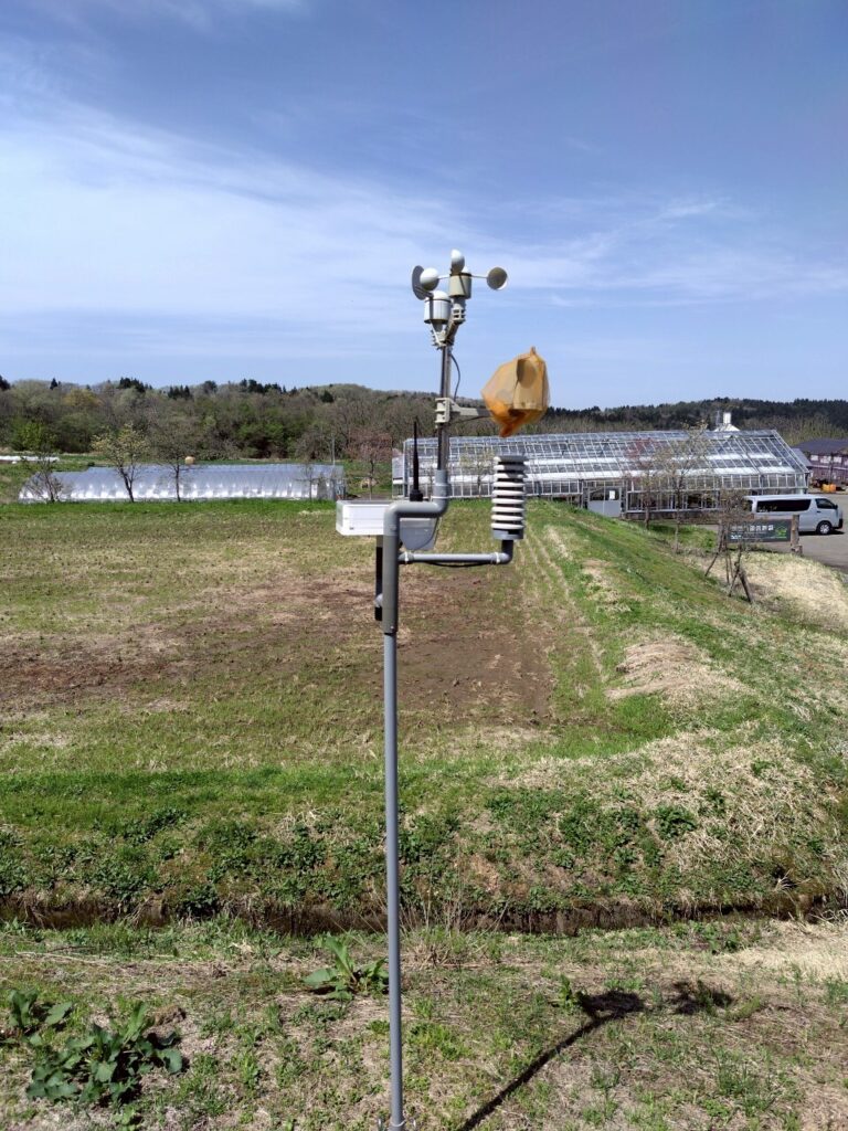 農地に設置された気象センサー。気温、風速計、湿度センサーなどを備え、周囲にはビニールハウスと農作業用の畑が広がっている。